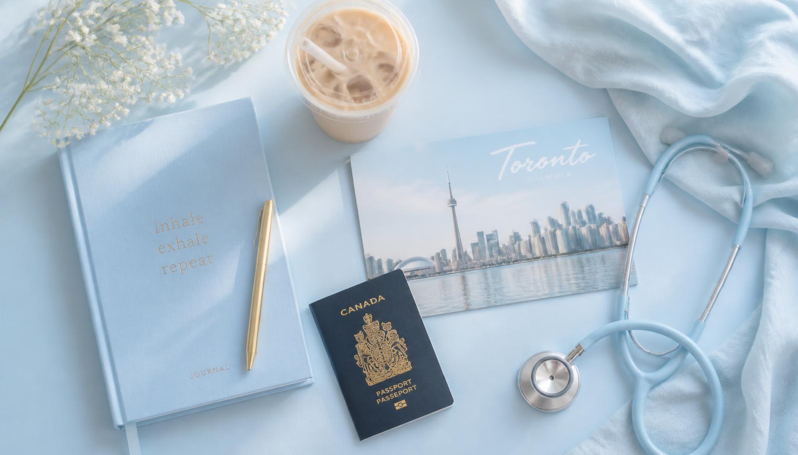 Baby blue flat lay with a journal, iced coffee, Toronto postcard, passport and stethoscope